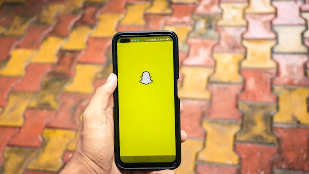 A person holds a smartphone displaying the Snapchat logo against a colorful brick background.