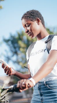 African American woman checking her phone while riding a bicycle for delivery outdoors.