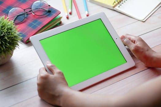 Close-up of hands holding a tablet with green screen, ideal for mockups. Surrounded by school supplies.