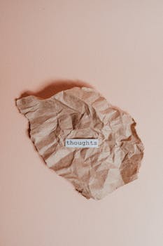 A crumpled brown paper on a pink background with the word 'thoughts' in focus.